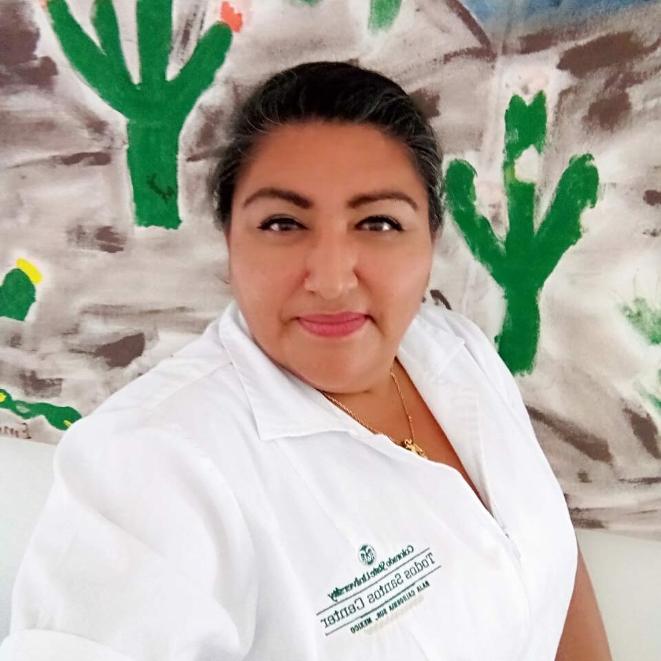Woman in a white shirt with the CSU Todos Santos logo in front of a wall of painted cacti.