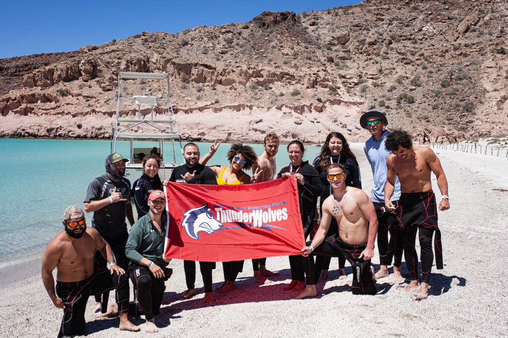 A group holds a red CSU Pueblo banner on a beach.