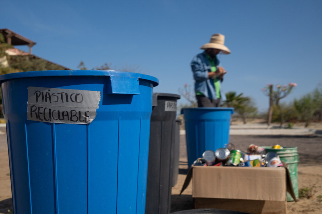 A man stands in the background with a blue trash can that says plastic recycling in the foreground.
